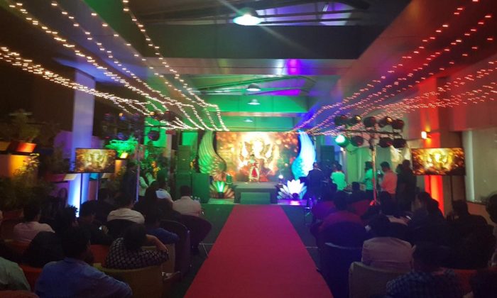 Indoor stage with a large illustrated deity/goddess backdrop, red carpet aisle and fairy-light drapery, seated audience; no legible branding.