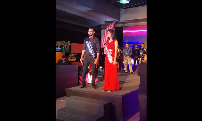 Two people on a small stage wearing pageant sashes reading "Mr" and "Miss ... 2018", posing together; no legible sponsor branding.