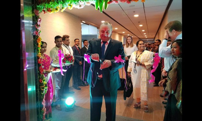A man in a suit cutting a ribbon under a floral arch in an office corridor, staff lined up on both sides; company signage not legible.