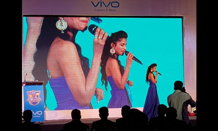 A female singer performing on the Vivo Champions Night stage in front of a teal LED screen showing her image.