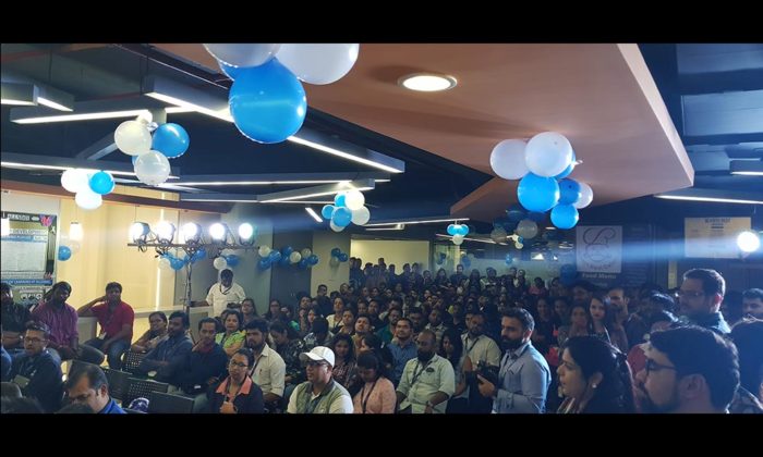 Wide shot of a large seated office audience under blue and white balloon decorations.