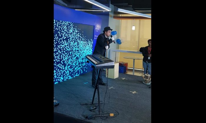 A performer in a hat holding a microphone/megaphone prop at a keyboard on a small stage with blue LED dot-pattern backdrop, part of the same office event.