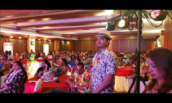 Audience wearing Hawaiian shirts and floral leis seated at an indoor event with a tropical backdrop.