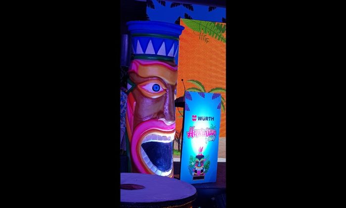 Man in a Hawaiian shirt and hat standing near the stage at a Hawaiian-themed party under colorful lighting.