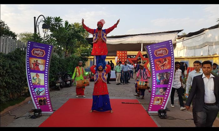 Bhangra performers on stilts in traditional Punjabi costume greet guests on a red carpet entrance flanked by purple "Concentrix" film-reel banners.