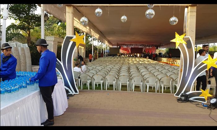 Empty canopy-covered outdoor seating area with rows of white chairs, hanging disco balls and gold star pillar decor, awaiting a show.