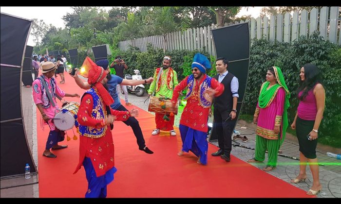 Bhangra dancers and dhol drummers performing on an outdoor red-carpet walkway lined by a wooden fence.