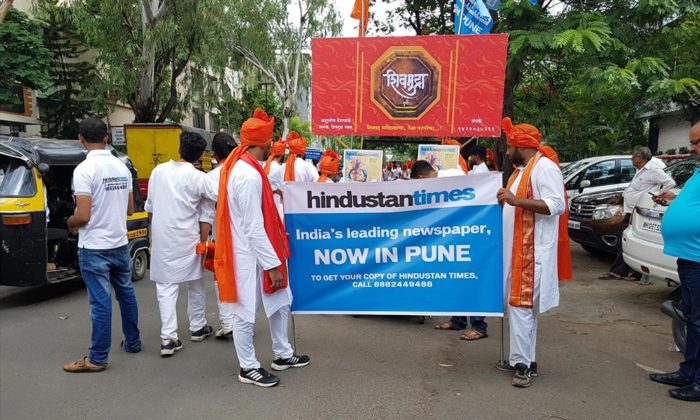 Street procession of men in saffron turbans and white kurtas carrying a red banner promoting Hindustan Times "Now in Pune".