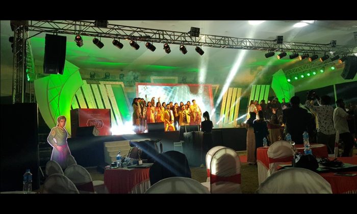 Dance group performing on an illuminated stage under a tented ceiling with green and gold lighting; a cardboard cutout figure stands at the stage edge.