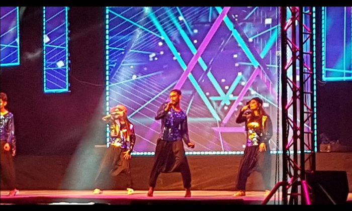 Performers in sequined jackets and harem pants singing and dancing on stage against a blue geometric LED backdrop, part of the same indoor gala.