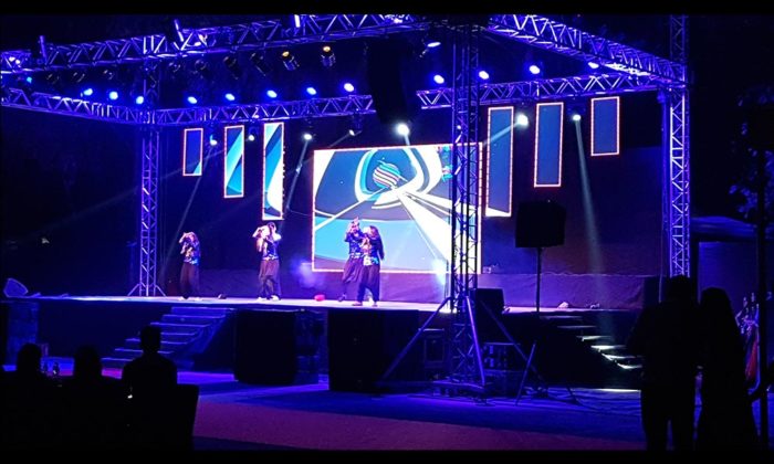 Dancers performing on an open-air truss stage lit in blue with a circular swirl-logo graphic on the LED screen, trees visible behind.