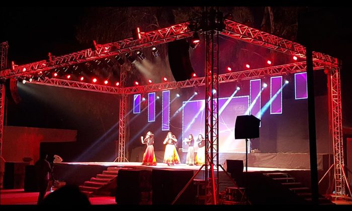 Three performers in folk attire singing on the same open-air truss stage lit in red.