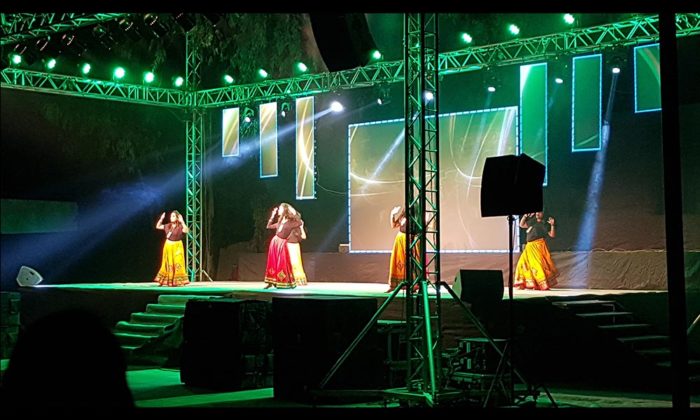 Four dancers in yellow and red folk skirts performing on the same open-air truss stage lit in green.
