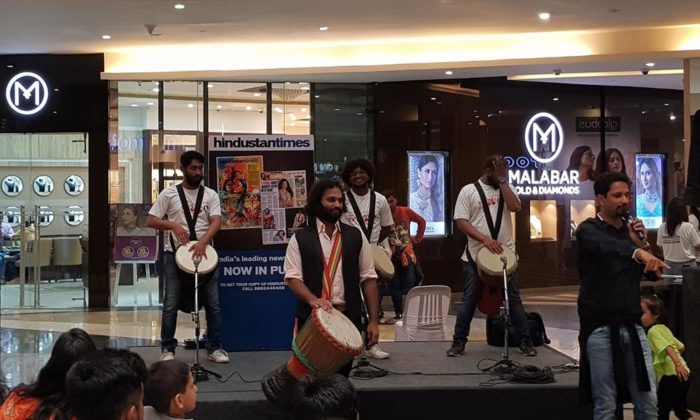 Djembe drum circle performers on a small stage in a shopping mall, Hindustan Times banner and Malabar Gold & Diamonds storefront visible behind.