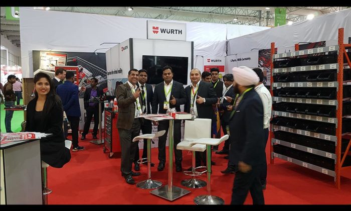 Staff and visitors posing at a "WÜRTH" branded trade show booth with tool and parts shelving on display, red-carpeted aisle.