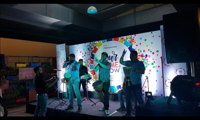 Drum circle performers on an indoor stage in front of a colorful polka-dot Concentrix-branded backdrop reading "Our [...] is Now".