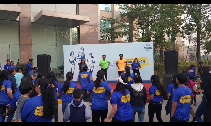 Outdoor stage warm-up session with instructors in orange and green, "Allstate India" and Walkathon banner behind, crowd in blue event t-shirts.