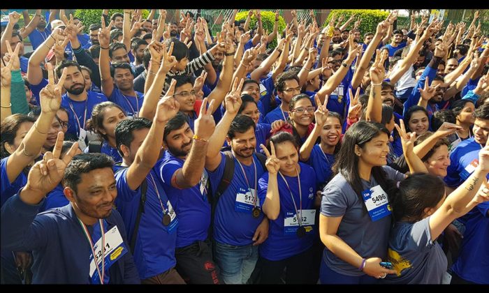 Large group of walkathon participants in blue t-shirts posing outdoors with peace signs and race bibs.