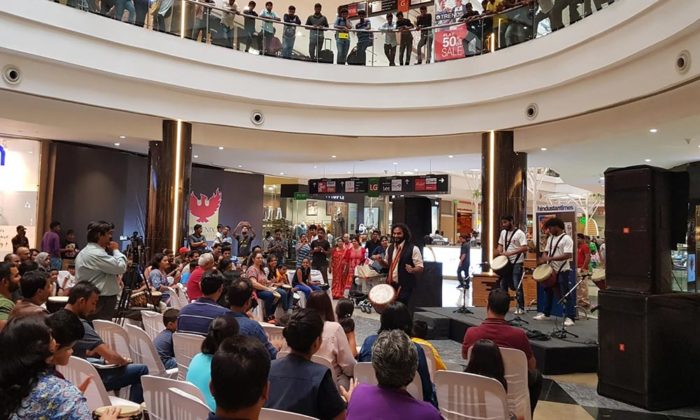 Wide view of a mall atrium with a seated crowd watching a drum circle performance on a stage bearing Hindustan Times signage.