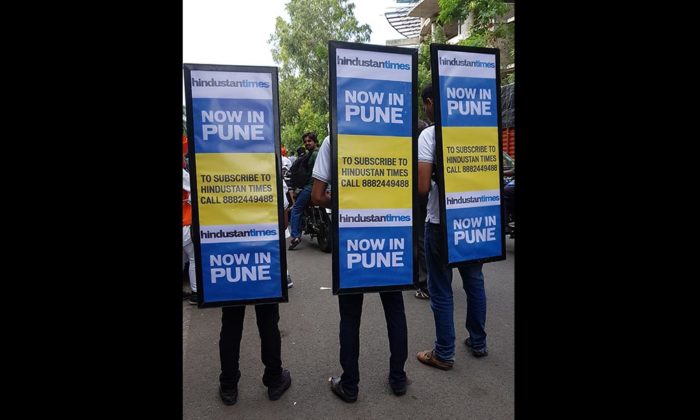Three people wearing sandwich-board costumes printed "Hindustan Times Now in Pune" with a subscription phone number, standing on a street.