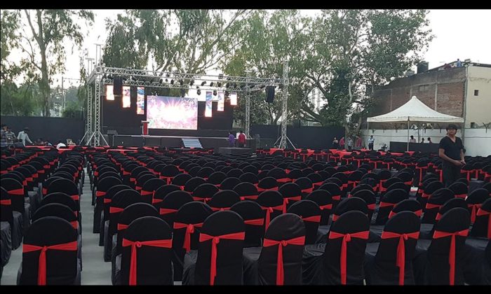 Empty outdoor evening event setup with rows of black chairs bearing red sashes facing a lit stage screen, marquee tent visible to the side; no legible branding.