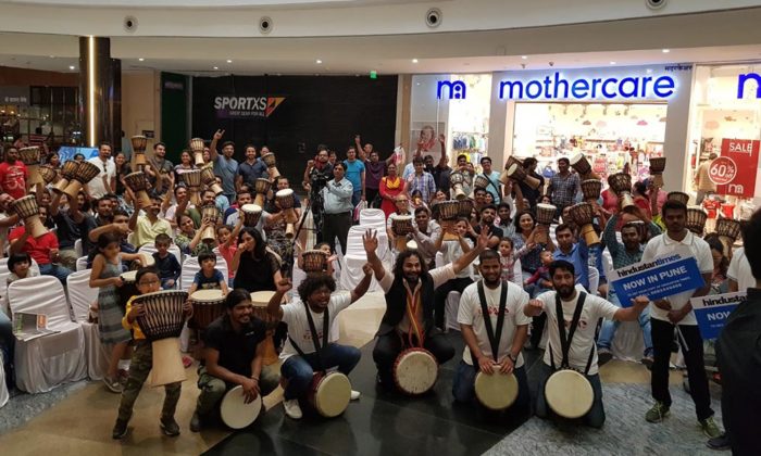 Large group photo of drummers, children and adults holding "Hindustan Times Now in Pune" placards near Mothercare and SportXS storefronts in a mall.