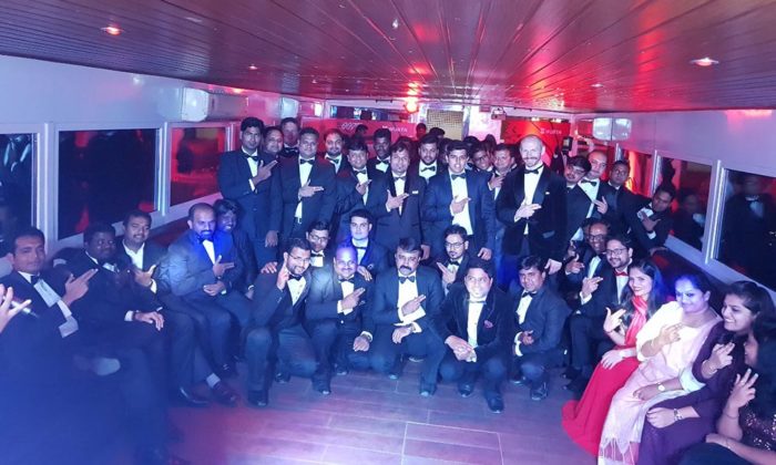 Large group of men in black-tie formal wear posing for a photo inside a boat or yacht cabin lit in blue and red; no legible branding.